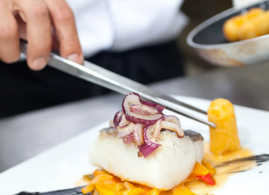 Plato gourmet con pescado, cebolla roja y guarnición de vegetales, decorado con pinzas.