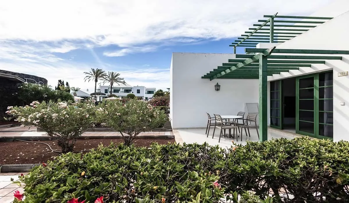 Casa blanca con pérgola, jardín y muebles de exterior bajo un cielo azul.