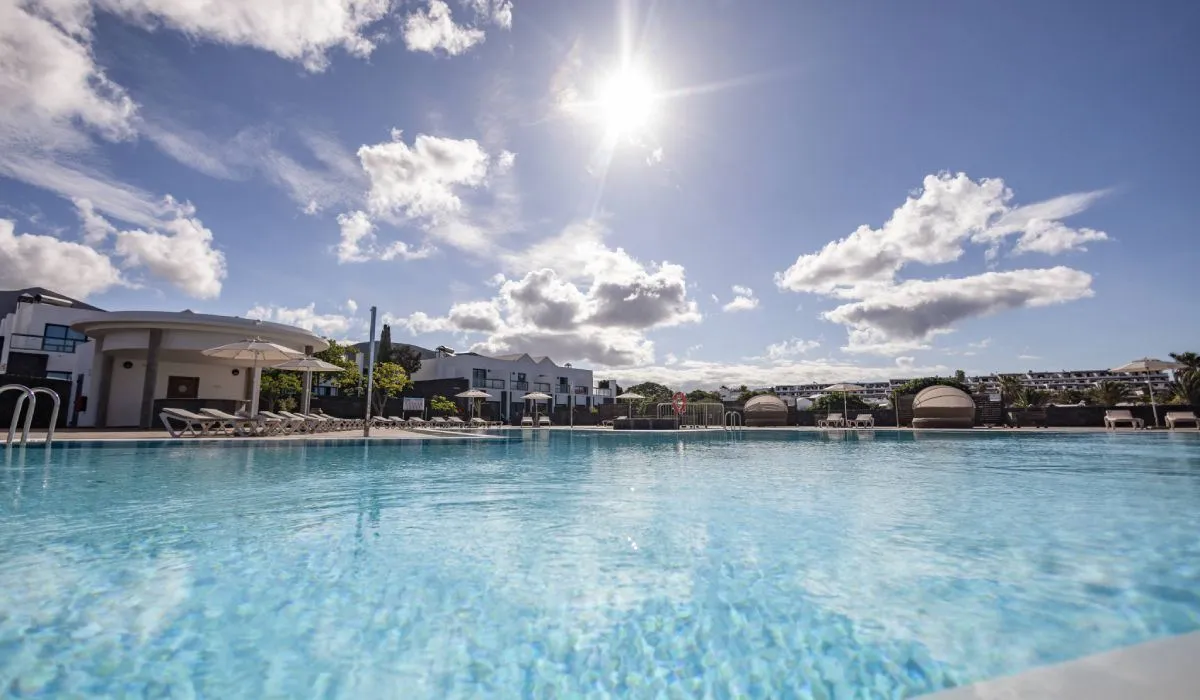 Piscina bajo un cielo azul con nubes esponjosas y el sol brillando.