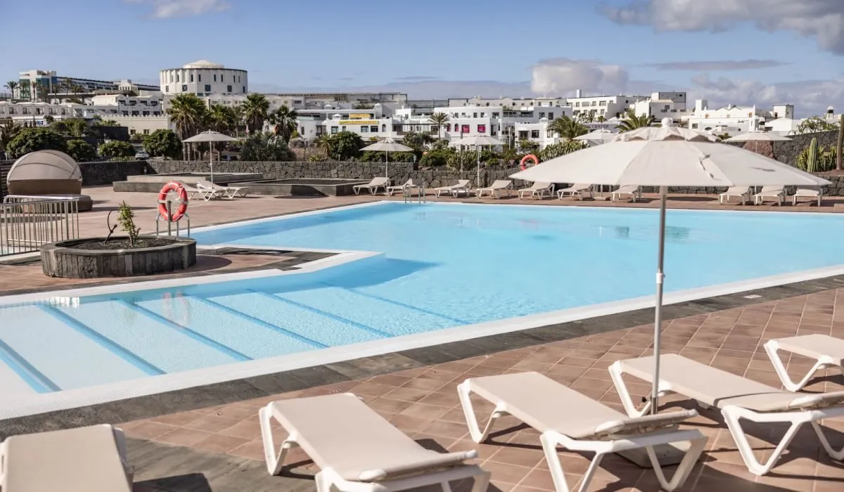 Piscina al aire libre con tumbonas y sombrilla, fondo de edificios y cielo.