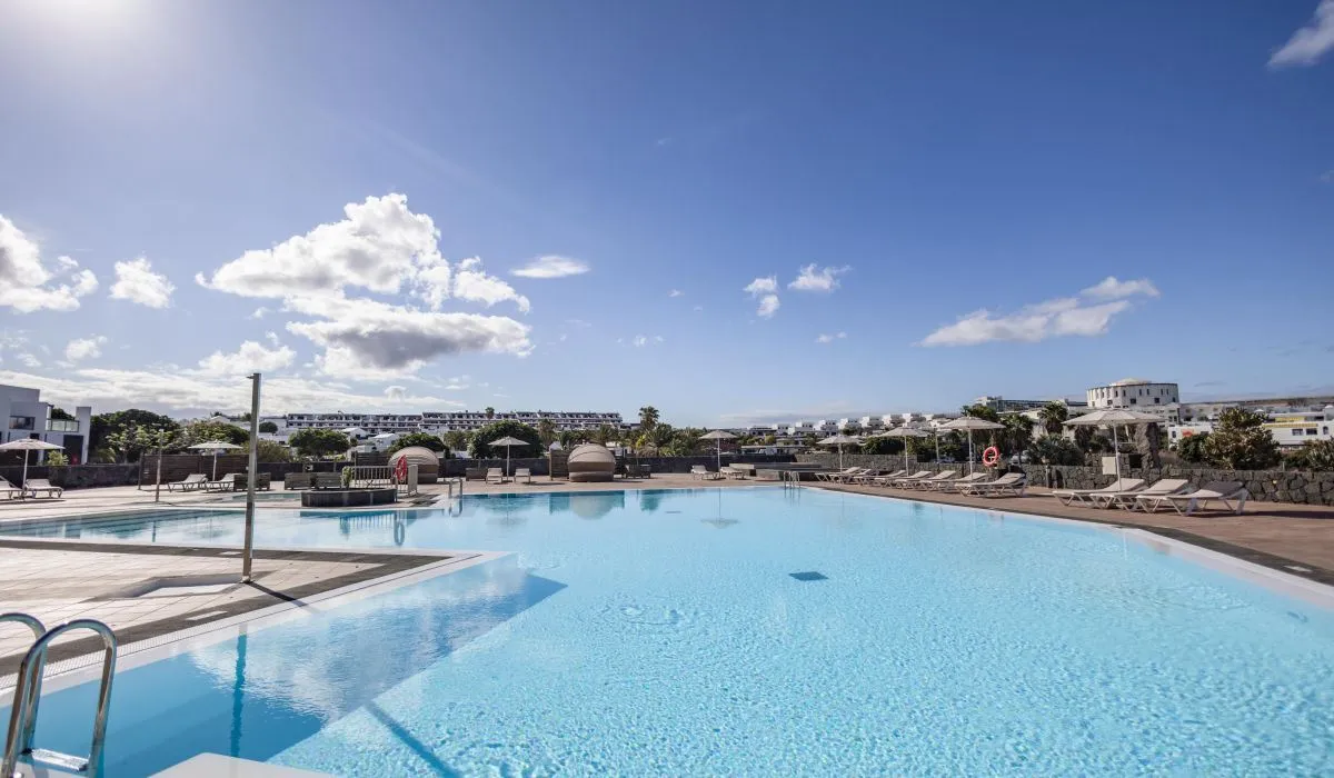 Piscina amplia con tumbonas, cielo azul y nubes dispersas alrededor.