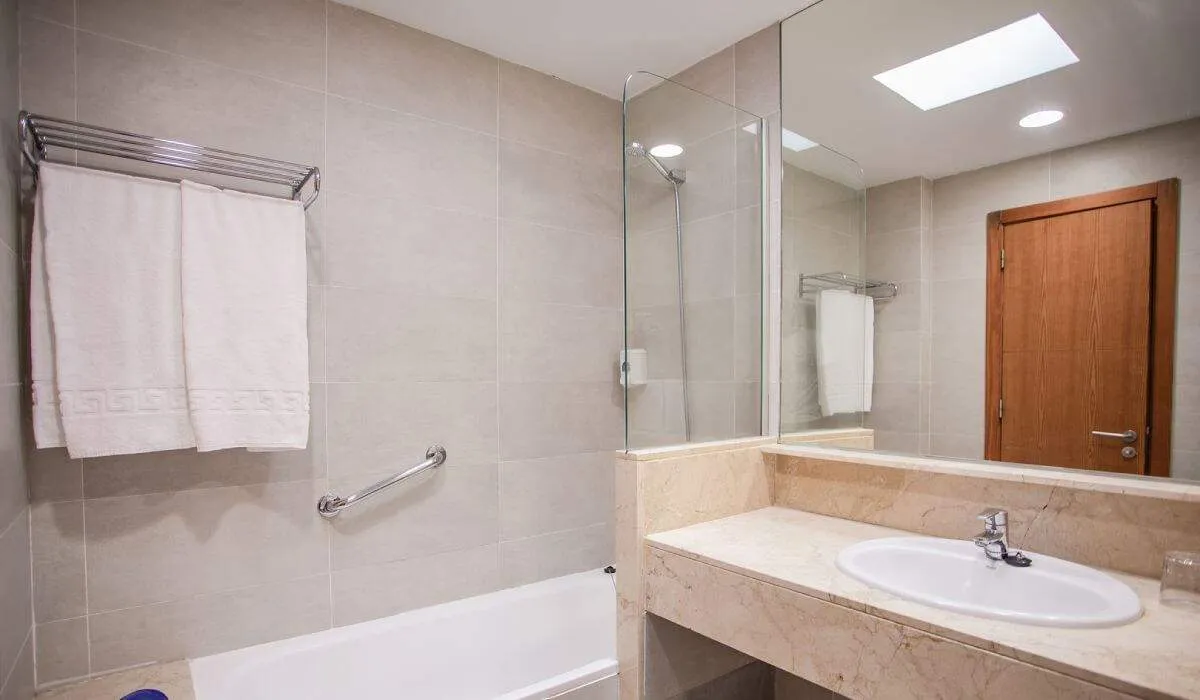 Baño moderno con lavabo, espejo, toallas y bañera. Puerta de madera al fondo.