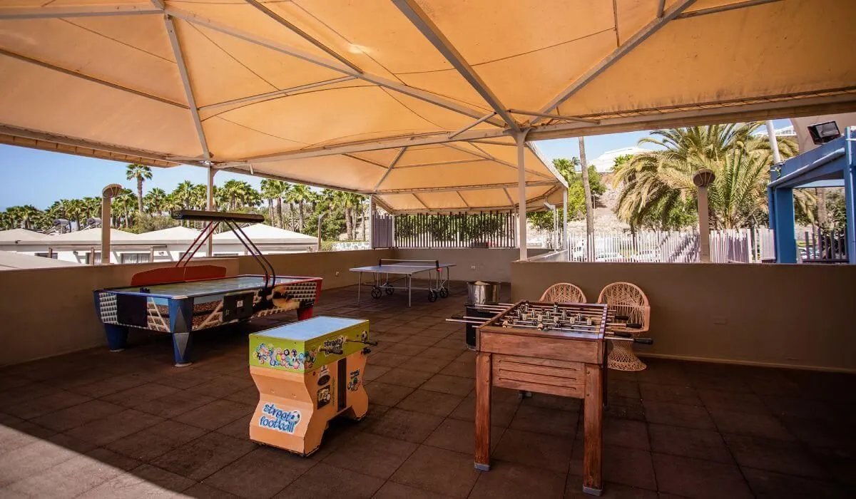 Terraza con mesas de juego y palmeras al fondo bajo techo de lona.
