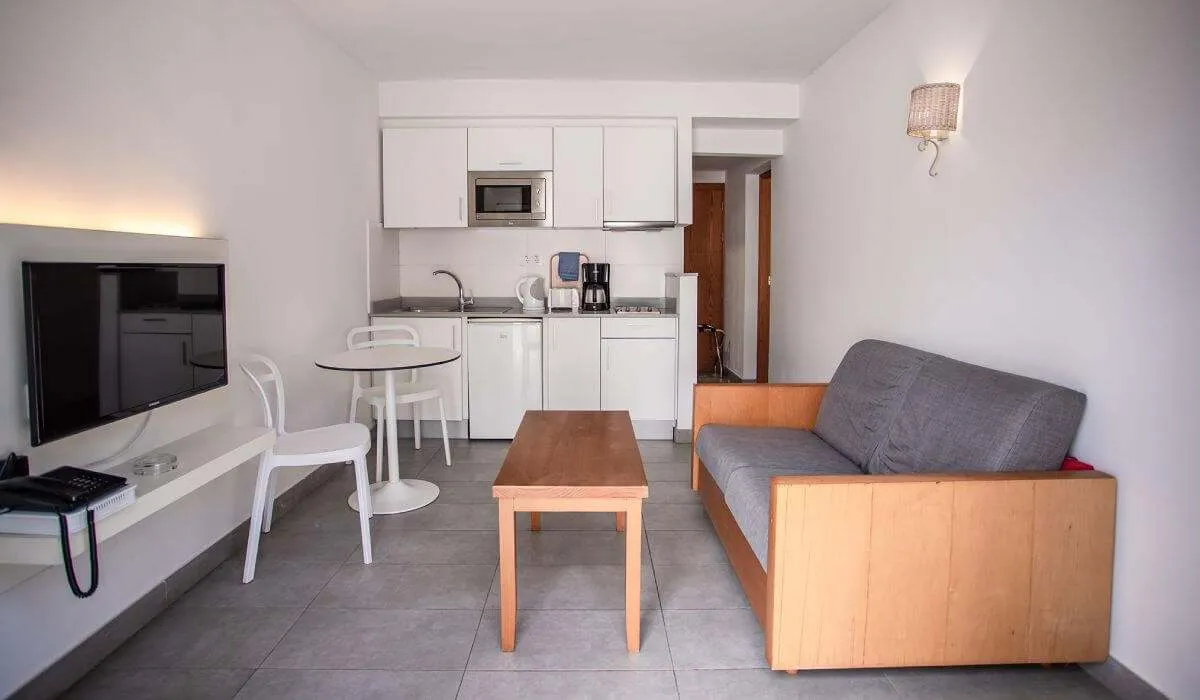 Sala con sofá, mesa, cocina pequeña y televisor montado en la pared.