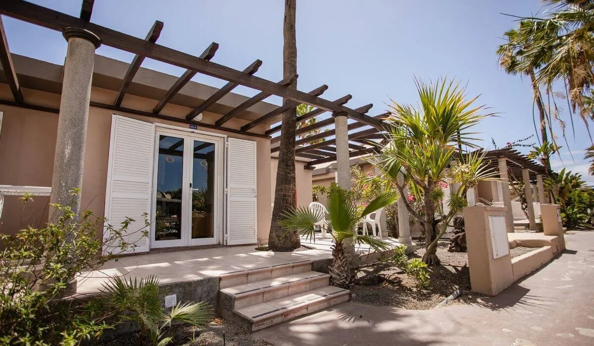 Casa con terraza, pérgola, plantas y sillas blancas en clima soleado.