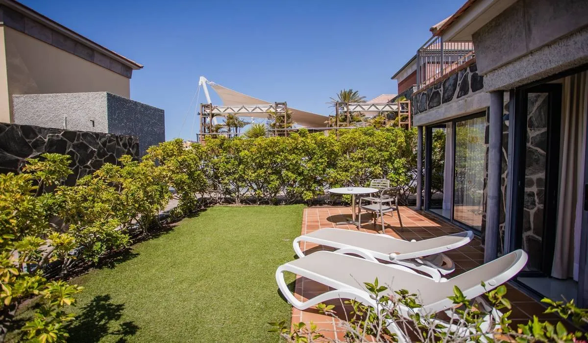 Terraza con tumbonas, mesa y sillas, rodeada de plantas y bajo cielo azul.
