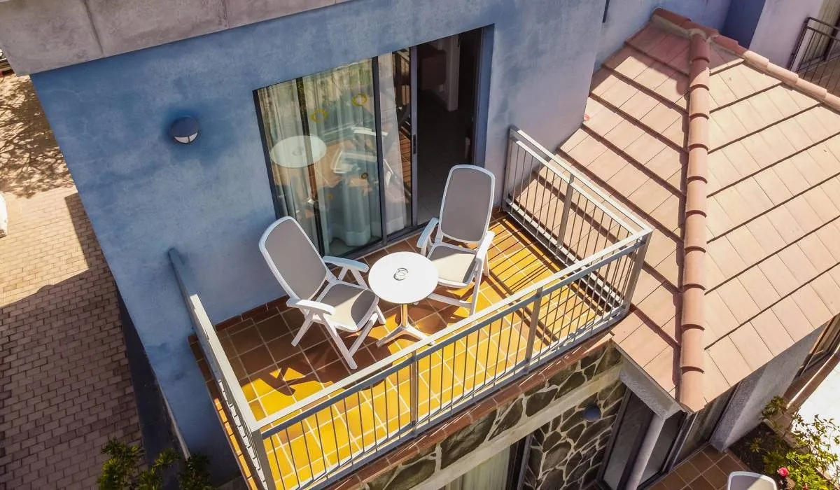 Balcón con dos sillas y mesa junto a edificio con paredes azules y tejado rojo.