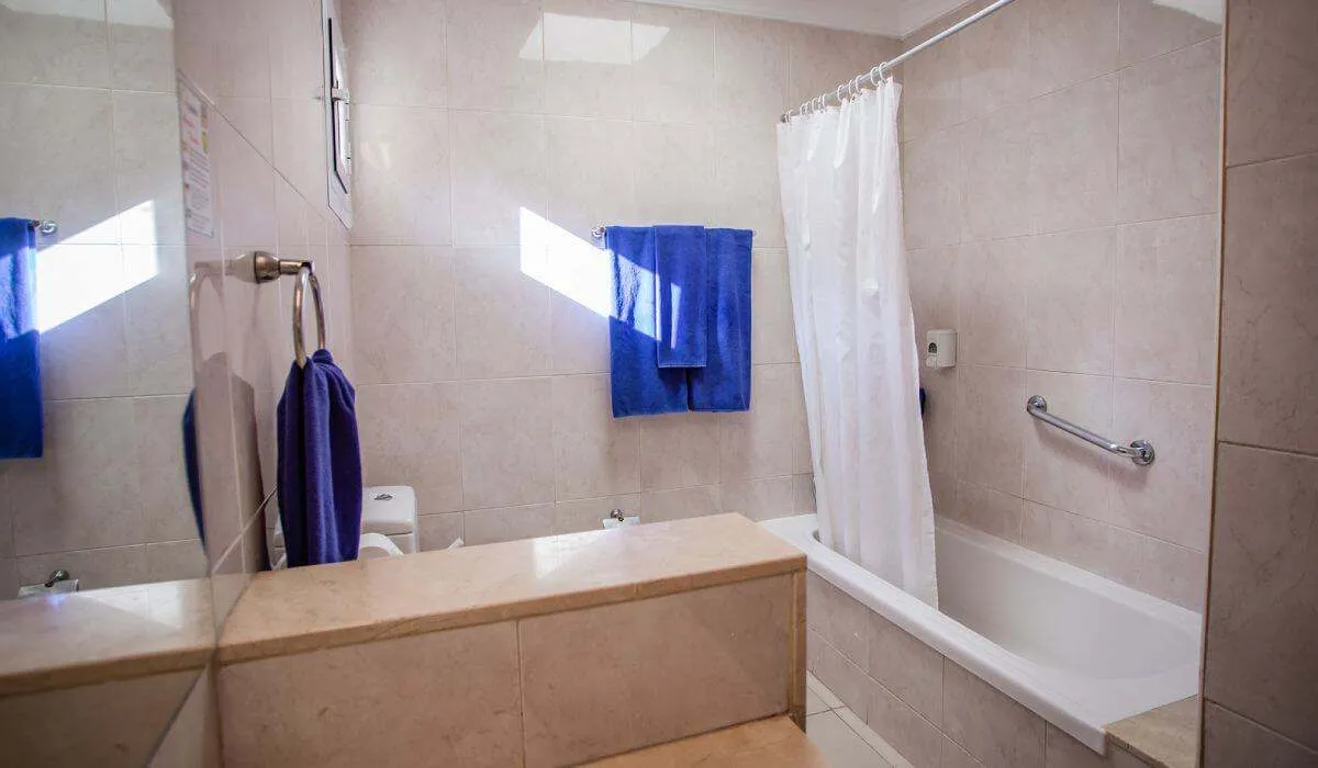 Baño con tina, cortina blanca y toallas azules en pared de azulejos claros.