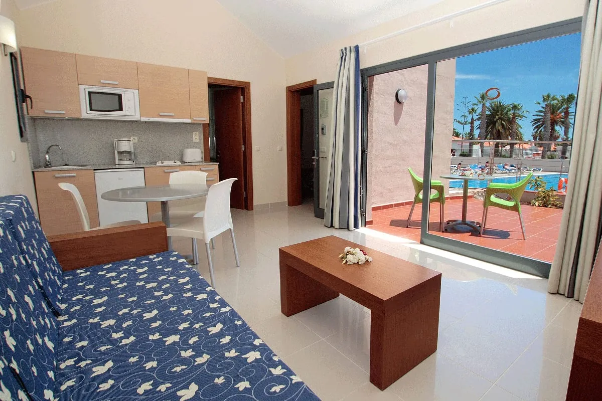 Sala con cocina, sofá, mesa y vista al exterior con piscina y palmeras.