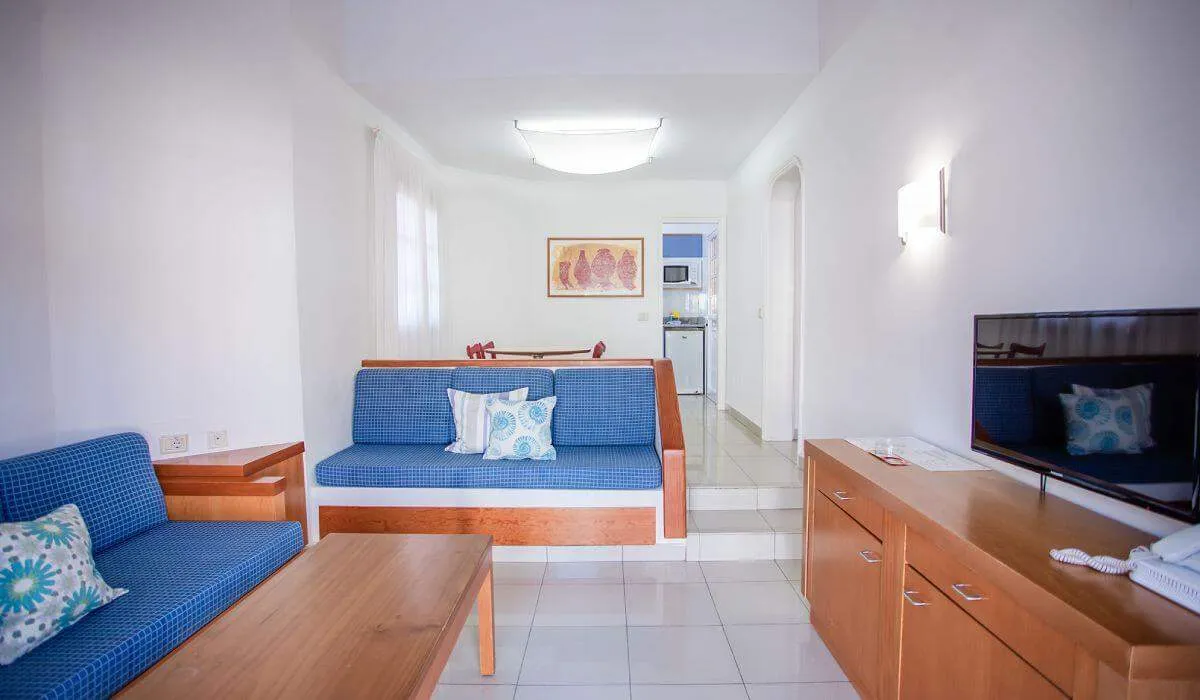 Sala de estar luminosa con sofá azul, TV y cocina al fondo.