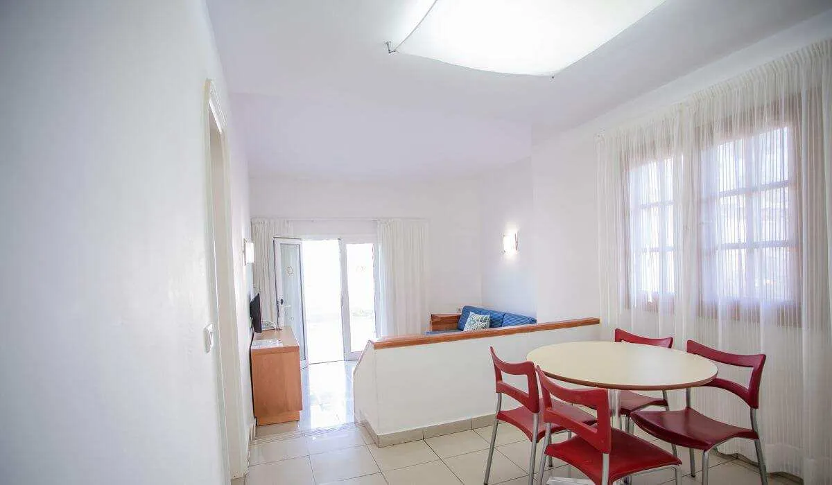 Sala moderna con mesa redonda y sillas rojas junto a ventanales blancos.