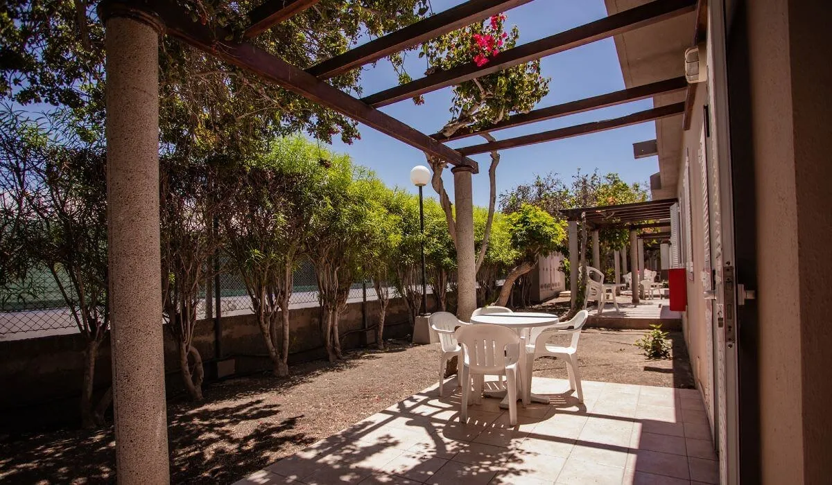 Patio exterior con mesa y sillas blancas bajo una pérgola de madera.