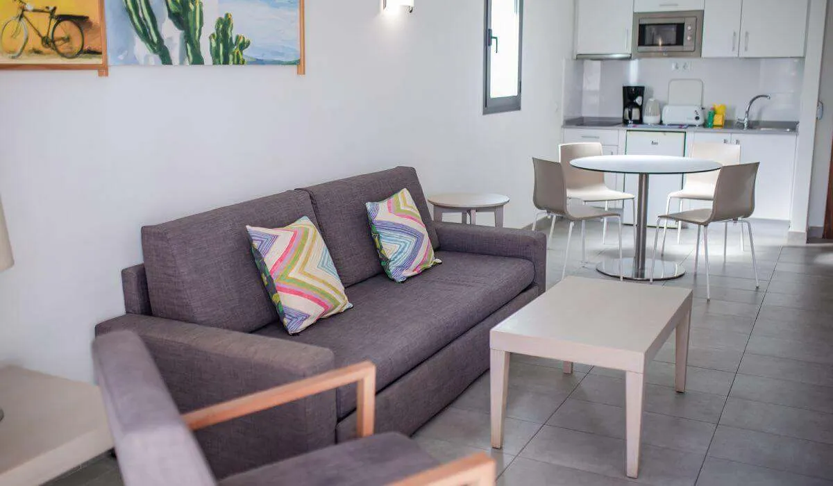 Sala con sofá gris, cojines coloridos y cocina integrada al fondo.