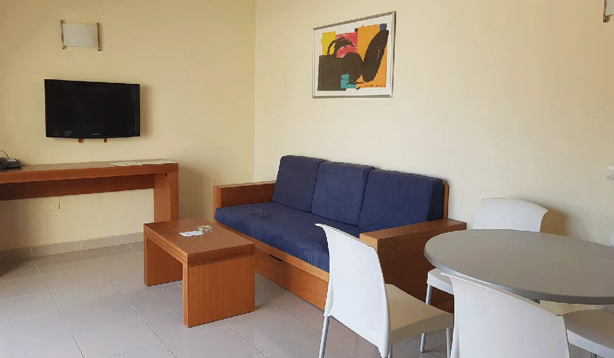 Sala minimalista con sofá azul, mesa, sillas, TV y cuadro en la pared.