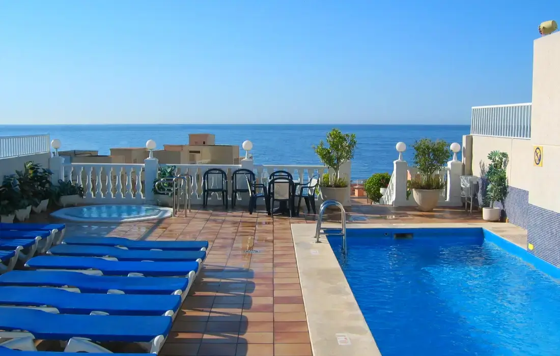 Piscina en azotea con vista al mar, rodeada de tumbonas y sillas.