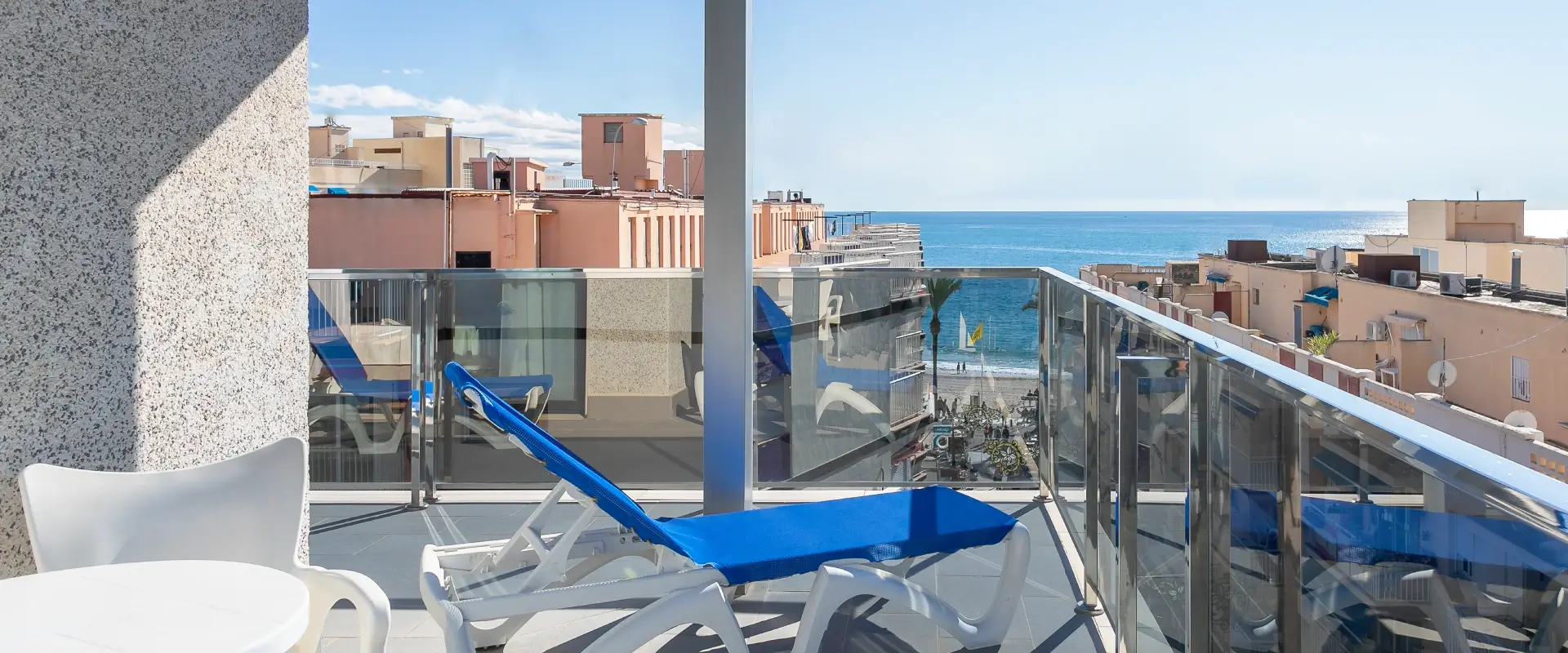 Terraza con vista al mar, tumbonas azules y edificios cercanos.