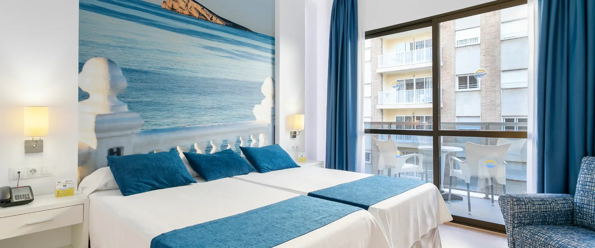 Habitación con dos camas, vista al balcón y decoración playera en la pared.