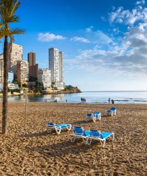 Playa con tumbonas, edificios al fondo, cielo azul y personas en el agua.