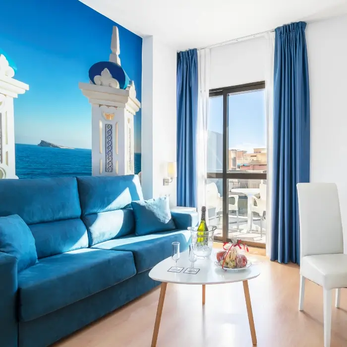 Sala moderna con sofá azul, mesa con vino y mural de paisaje marino.