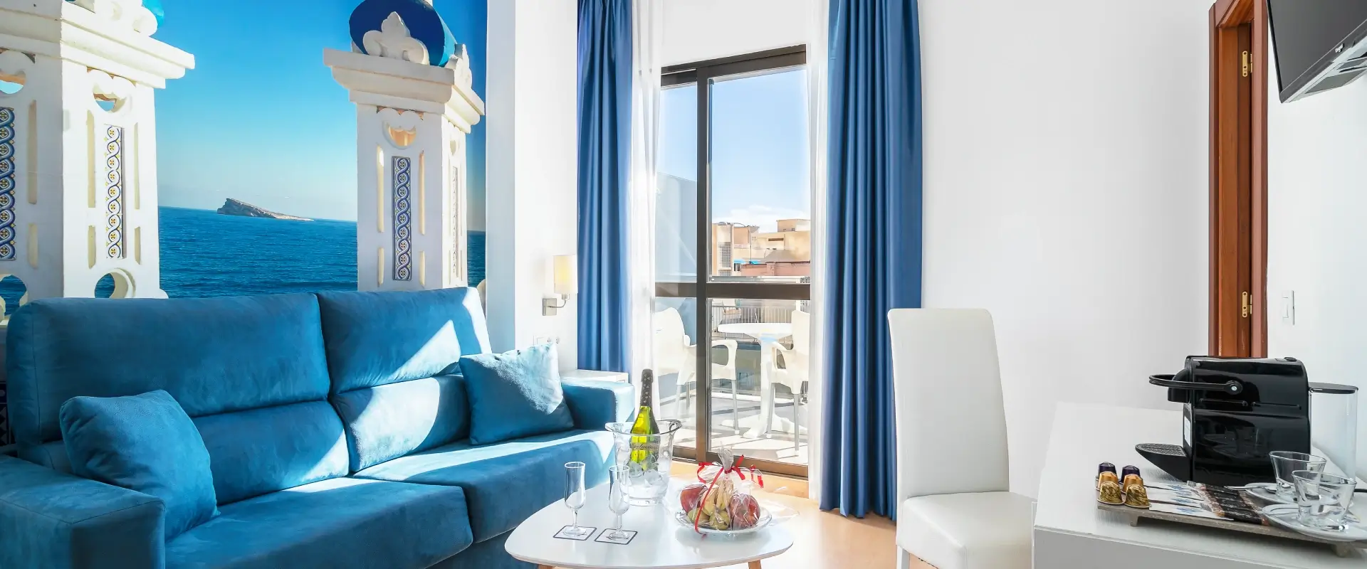 Sala luminosa con sofá azul, balcón y cafetera sobre mesa blanca.