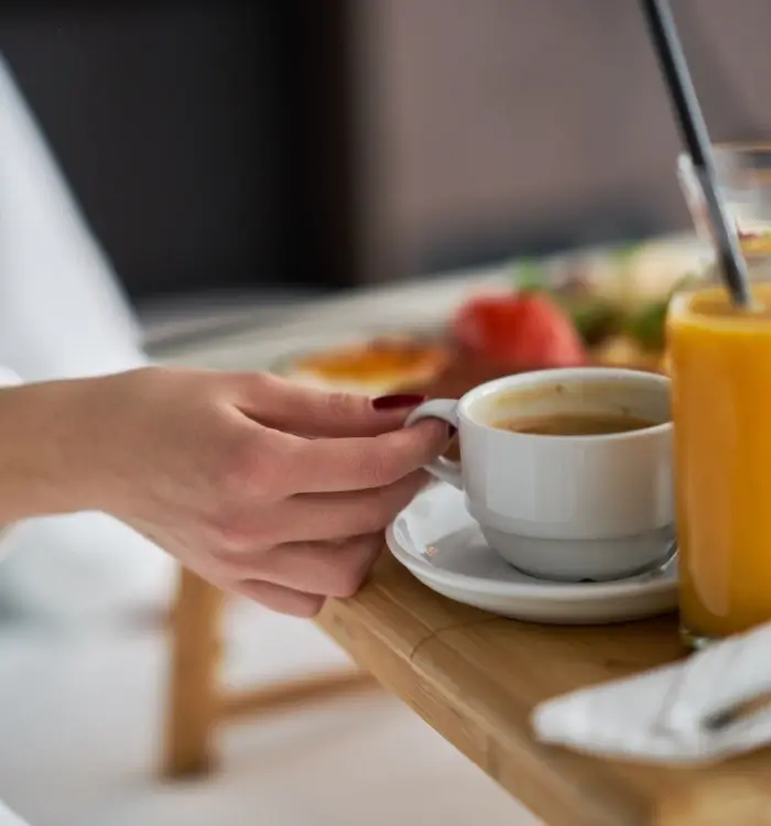 Mano sosteniendo taza de café junto a un vaso de jugo en una mesa de desayuno.