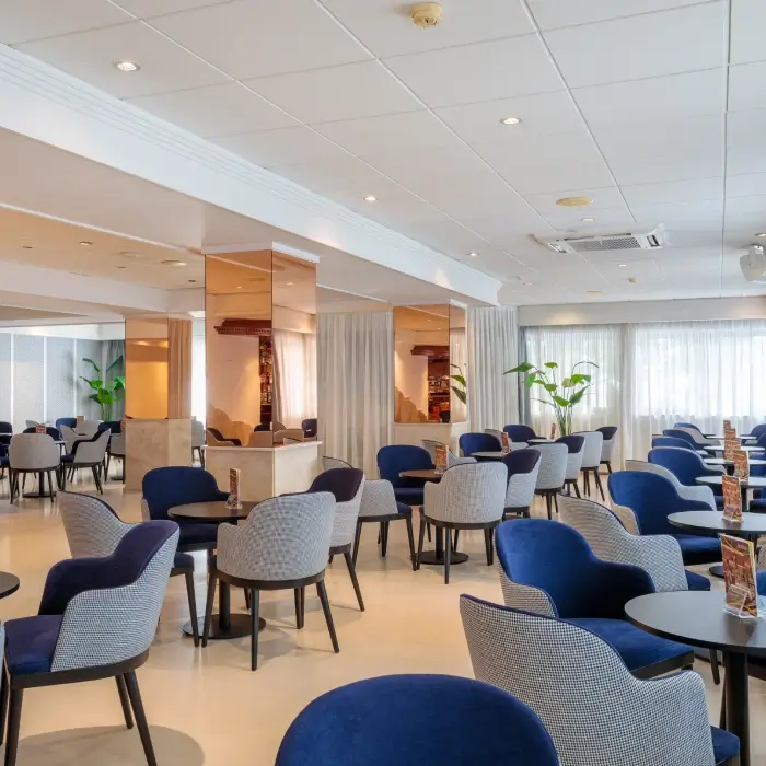 Sala de descanso moderna con sillas azules y grises, mesas redondas y plantas.