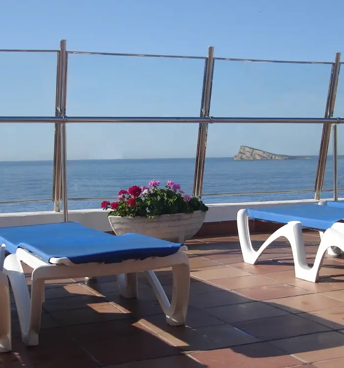 Terraza con tumbonas azules, flores y vista al mar.