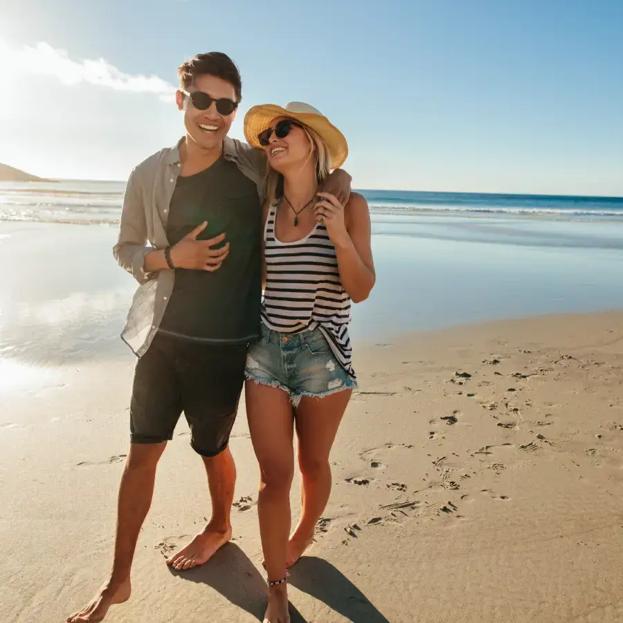 Pareja sonriente caminando descalza por la playa bajo un cielo azul.