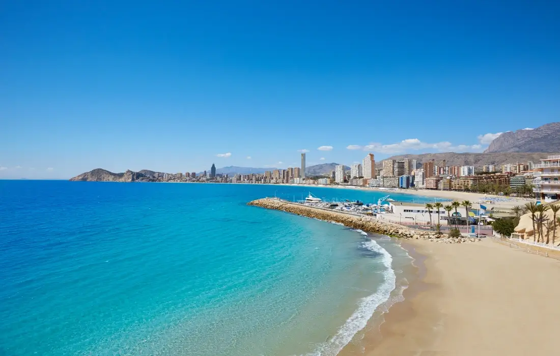 Playa con edificios y montañas al fondo bajo un cielo azul despejado.