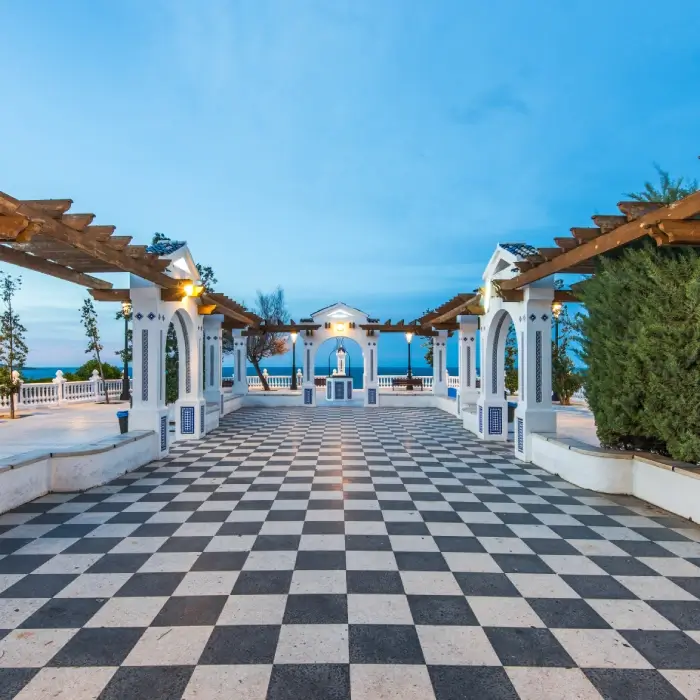 Plaza con suelo ajedrezado y pérgolas en un entorno costero al atardecer.