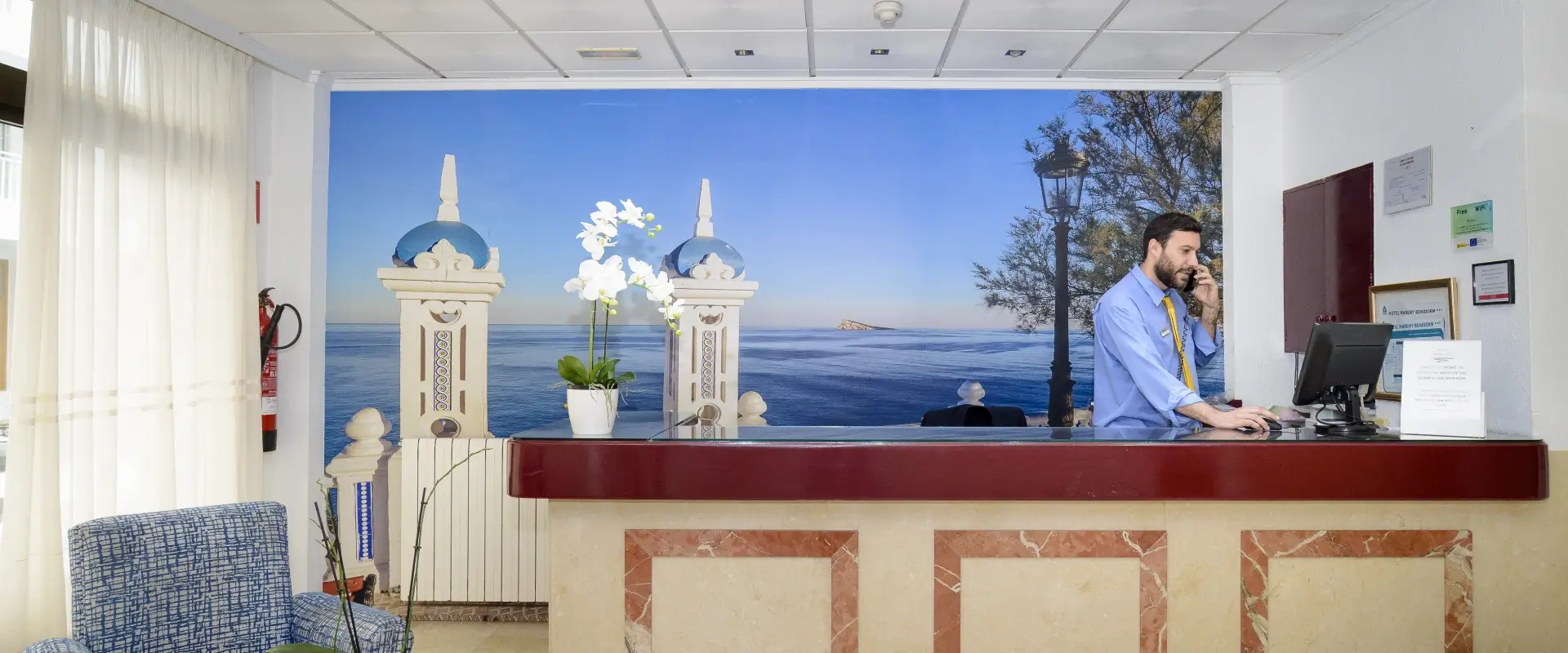 Recepción de un hotel con mural del mar y recepcionista al teléfono.