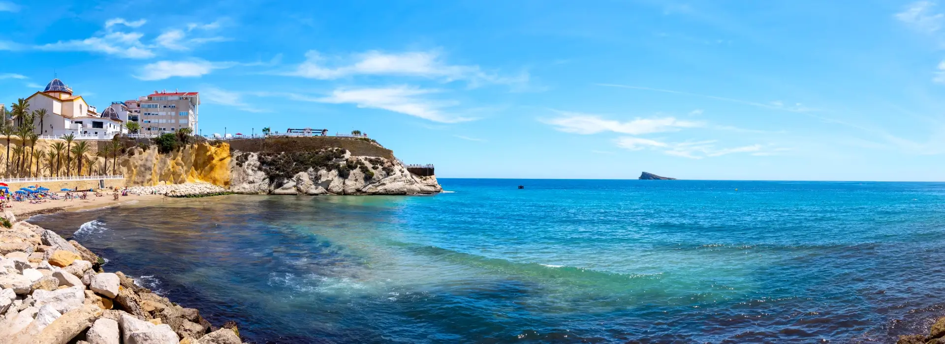 Playa con acantilado, edificaciones y mar azul bajo un cielo despejado.