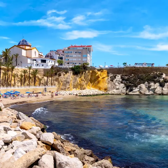 Playa con acantilados, edificios y palmeras bajo un cielo azul.