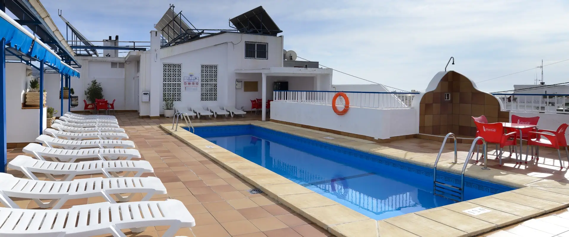 Piscina en azotea con tumbonas y mesas rojas bajo un cielo despejado.