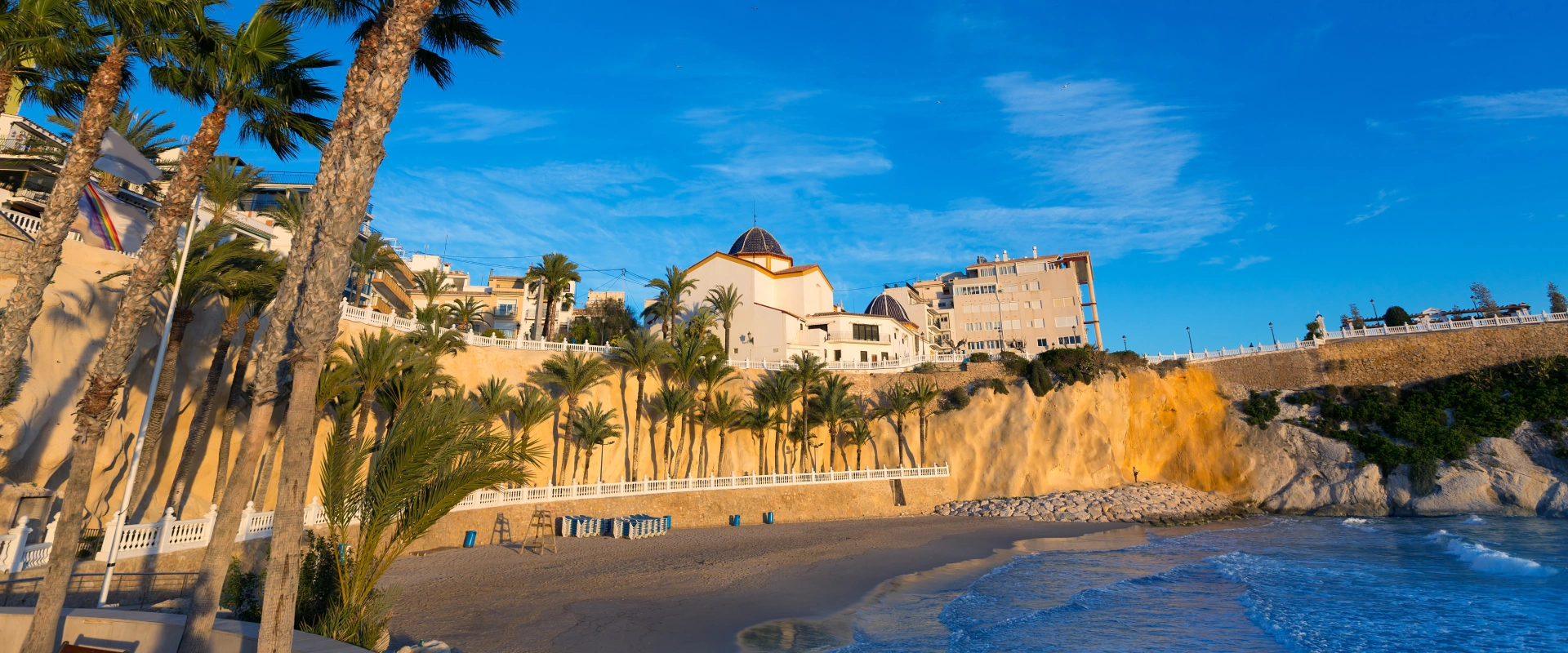 Playa con palmeras, acantilado y edificios al fondo bajo un cielo azul.
