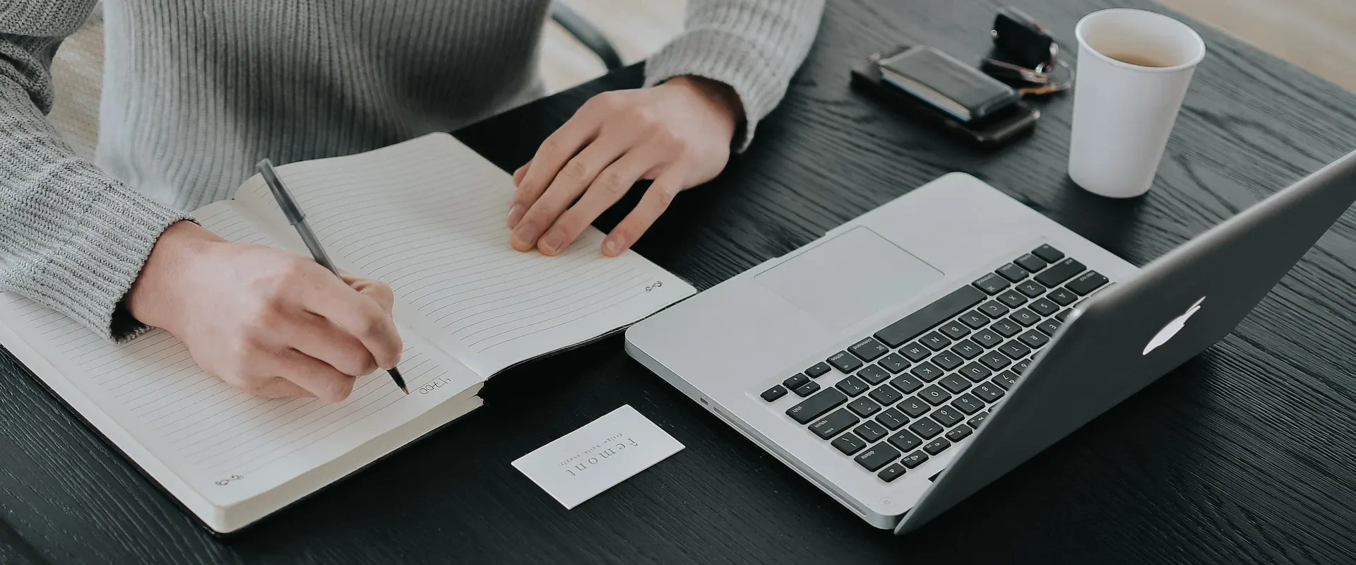 Persona tomando notas frente a una laptop con una bebida.