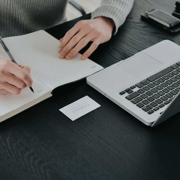 Persona escribiendo en cuaderno junto a laptop sobre escritorio negro.