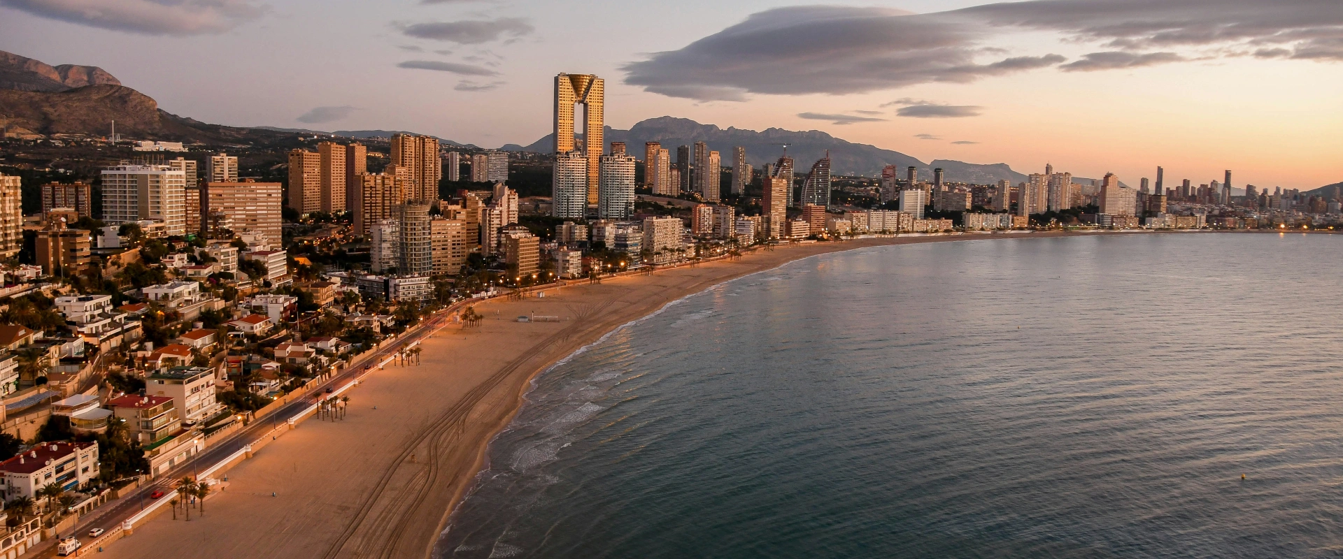 Playa y rascacielos al atardecer, con montañas al fondo y cielo parcialmente nublado.