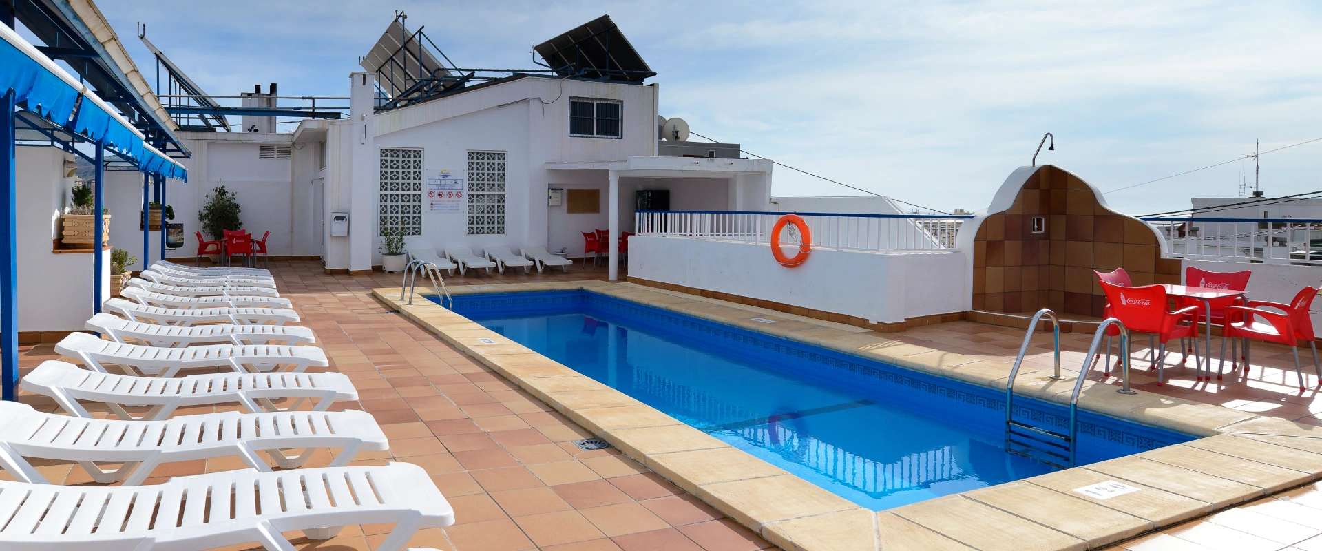 Piscina en azotea con tumbonas blancas y sillas rojas bajo cielo azul.