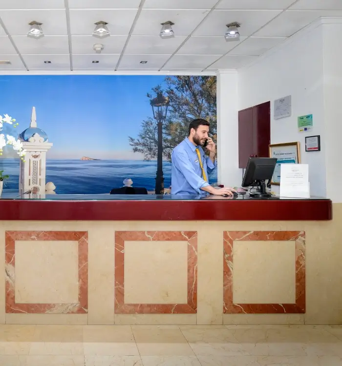Hombre atiende recepción de hotel con fondo de paisaje marítimo.