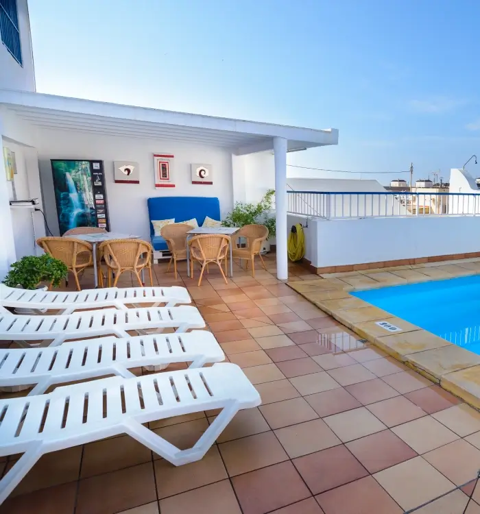 Terraza con piscina, tumbonas y mesa con sillas bajo un techo blanco.