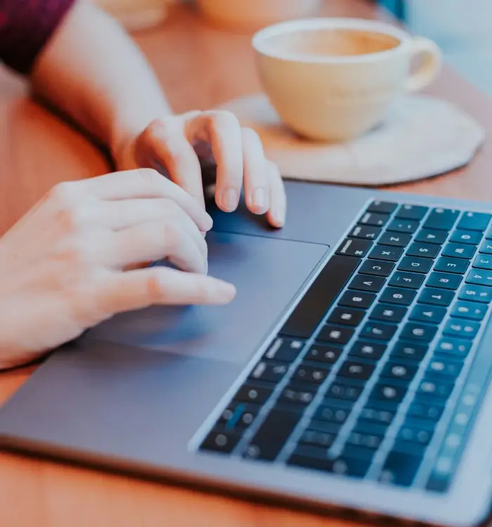 Manos usando una laptop con una taza de café al fondo.