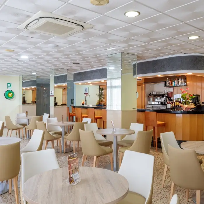 Cafetería moderna con mesas y sillas vacías, barra al fondo.