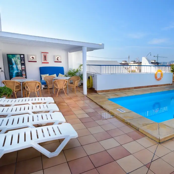 Terraza con piscina, tumbonas y mesa con sillas al lado de una pared blanca.