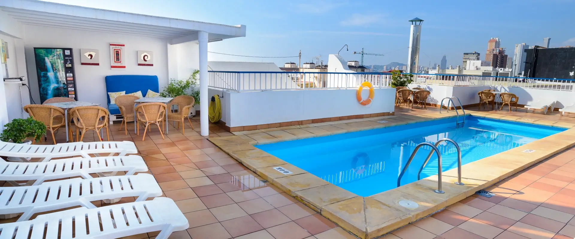 Terraza con piscina, tumbonas y mesas, rodeada de edificios urbanos.