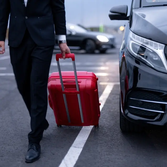 Hombre de traje con maleta roja camina junto a un auto negro en estacionamiento.