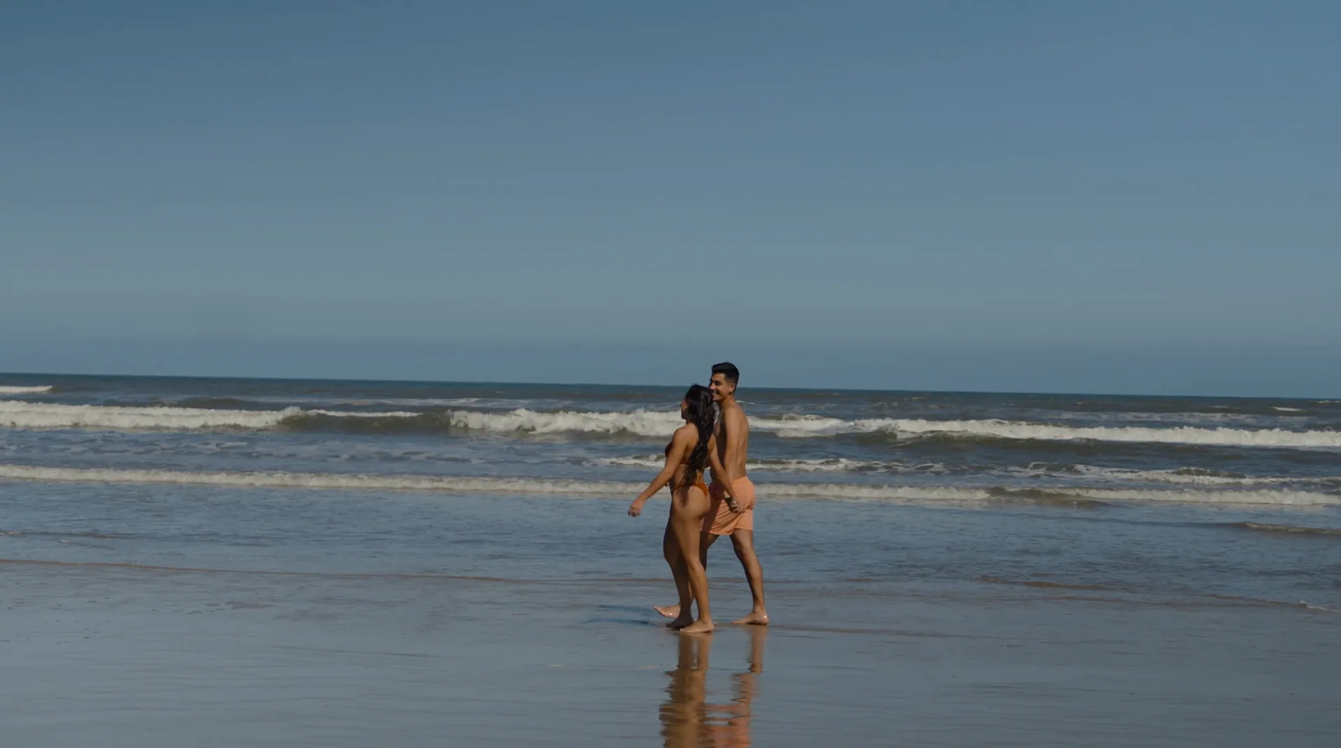 Pareja caminando por la orilla de una playa con el mar y cielo despejado de fondo.