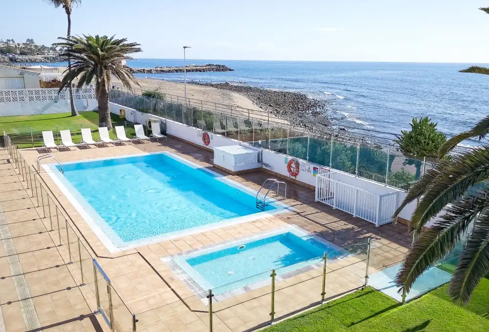 Piscina junto al mar con sillas reclinables y palmeras bajo un cielo despejado.