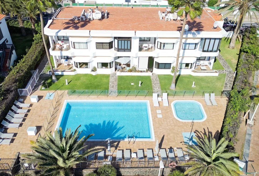 Edificio blanco con piscina y jard&iacute;n, rodeado de tumbonas y palmeras.
