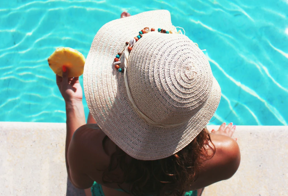 Mujer con sombrero de paja, comiendo piña, sentada al borde de una piscina azul.
